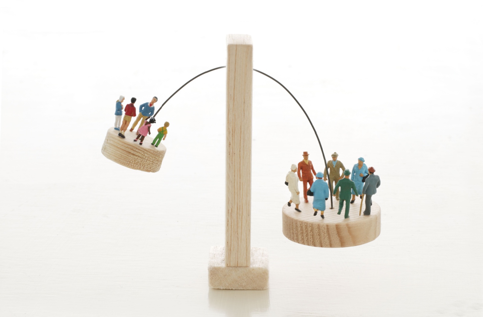 A balancing scale with miniature figures, showing fewer young people on one side and a larger group of older adults on the other, symbolizing the growing aging population